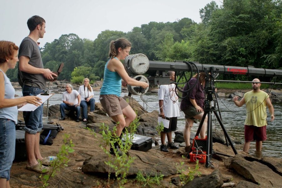 Final Acts film crew operates camera crane on rocky riverbank with cast and crew gathered around equipment