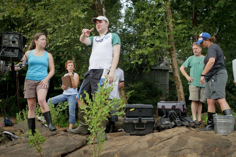 Director in cap gestures while standing on rock ledge with crew on outdoor film set in wooded area