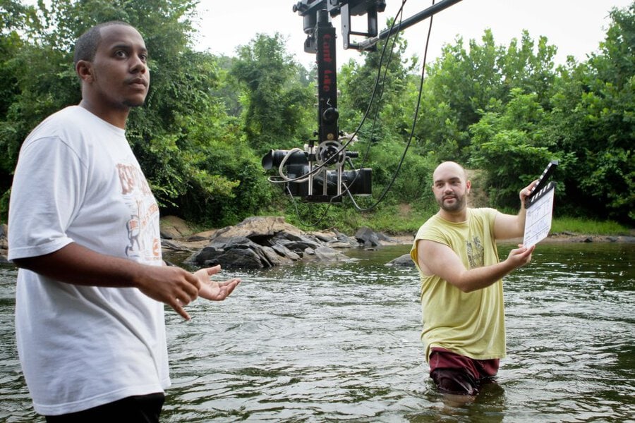 Two crew members standing in shallow river water during film production, camera rig overhead, lush Georgia trees behind
