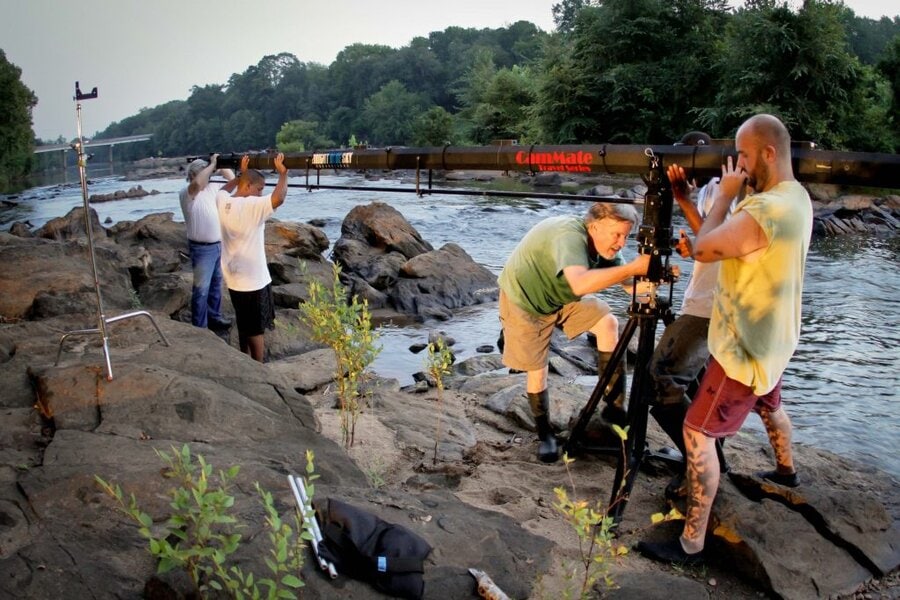 Film crew operates camera crane on rocky riverbank at sunset, crew members adjust rigging, trees in background