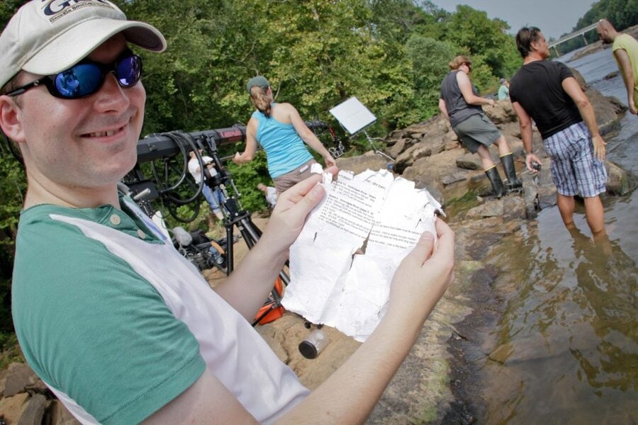 Crew member holding wet, tattered script pages beside river during Final Acts production in Macon, Georgia