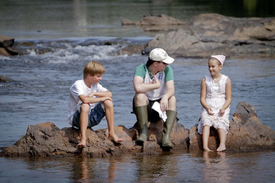 Two children and adult in white baseball cap sit on river rocks during Final Acts film shoot in Macon, Georgia