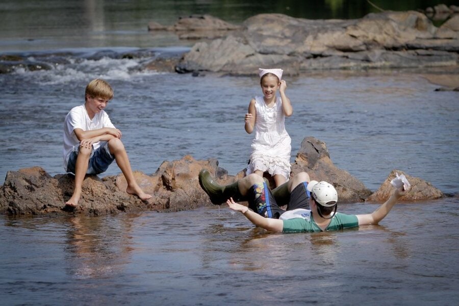 Three children play in a shallow river, one floating in an inner tube while two sit on muddy rocks