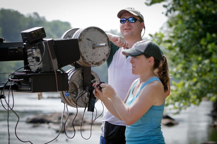 Man in white shirt and cap pointing while woman in blue tank top operates professional camera outdoors in Macon, GA