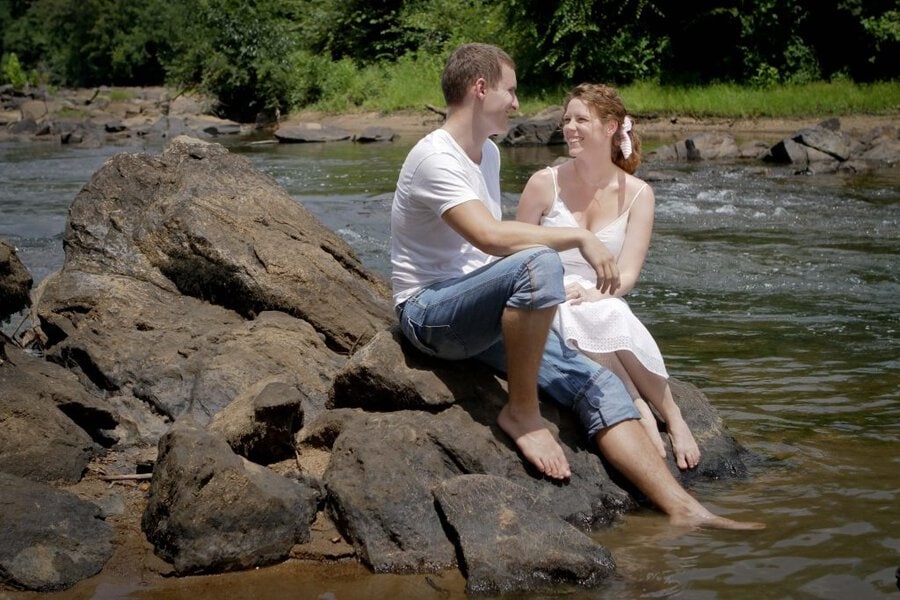Man and woman in white clothing sit on rocks with feet in flowing river, lush green trees in background