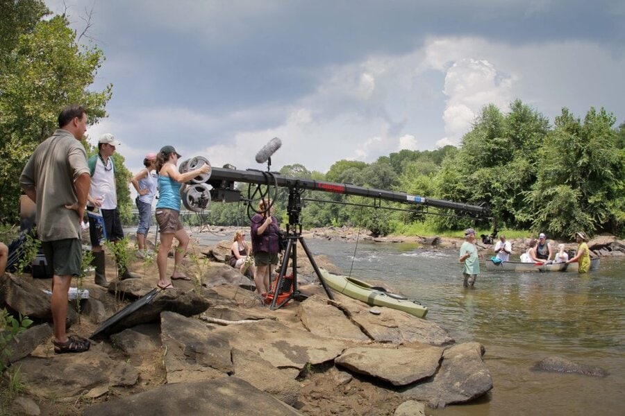 Film crew operating camera jib over rocky riverbank while kayakers float in background, Macon GA