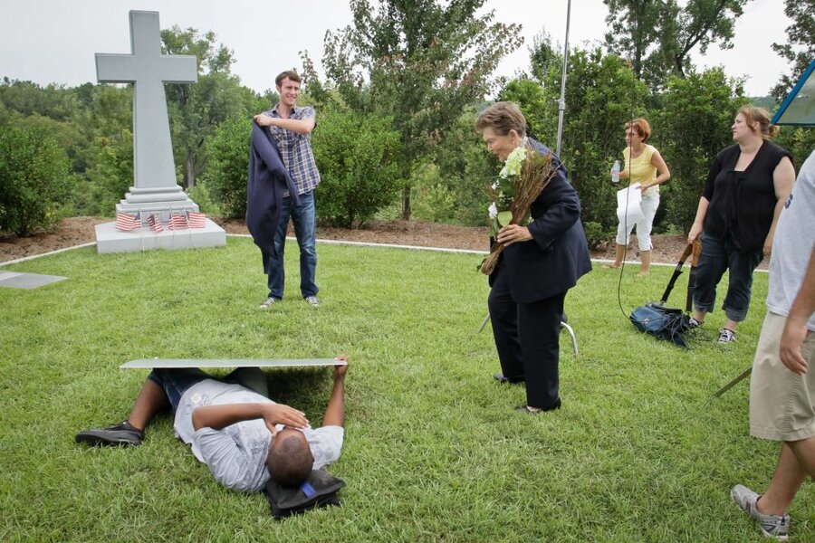 Actor lying on grass during funeral scene rehearsal while crew and cast members look on, large cross monument behind