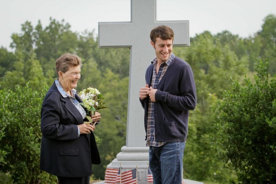 Two people share a moment at a white memorial cross in a cemetery, one holding white flowers, American flags at base