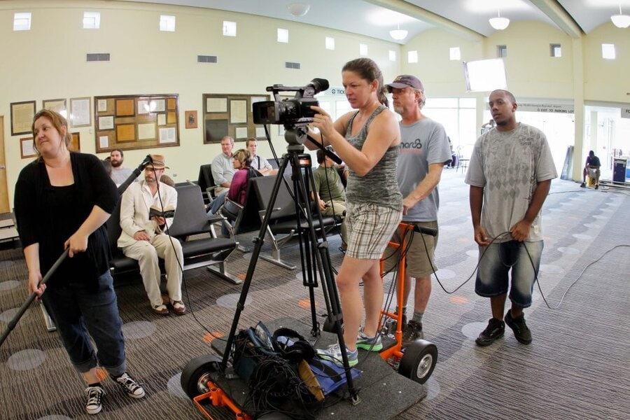 Film crew operating video camera on wheeled dolly in community center, audience seated behind