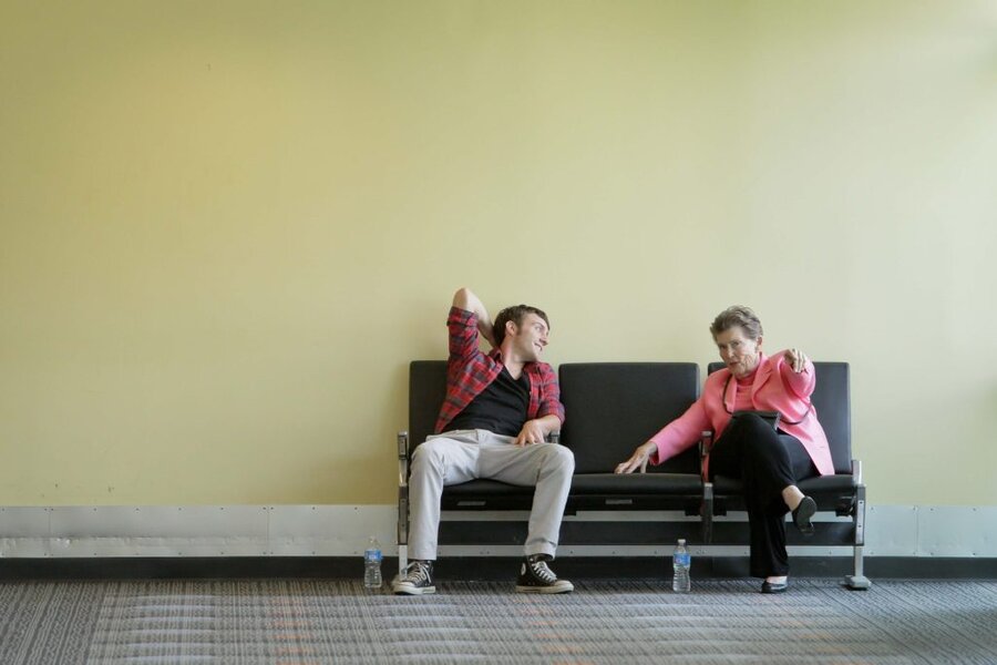 Two people seated on black couch against yellow wall, man in plaid stretching, woman in pink blazer pointing