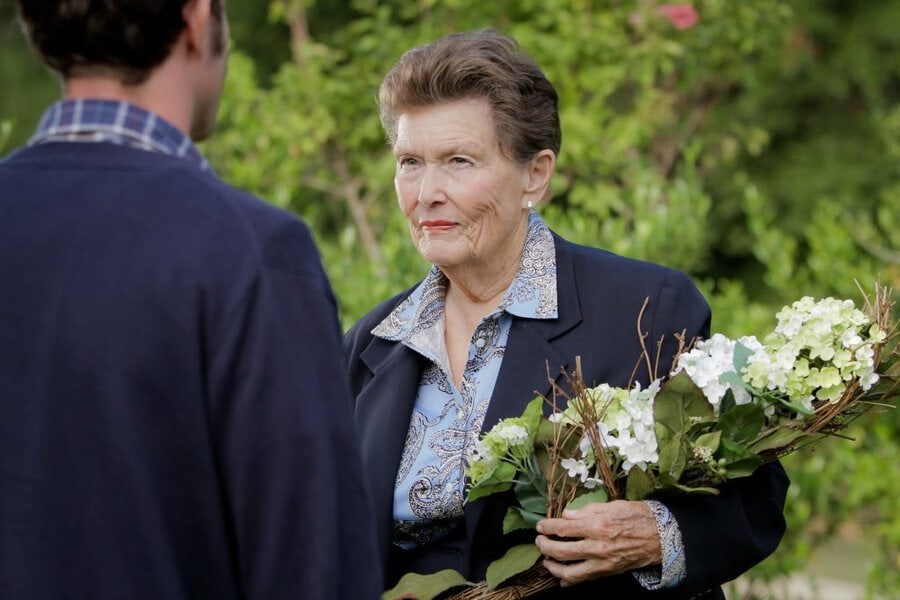 Older woman in black blazer holding white hydrangeas speaks with person in outdoor garden setting