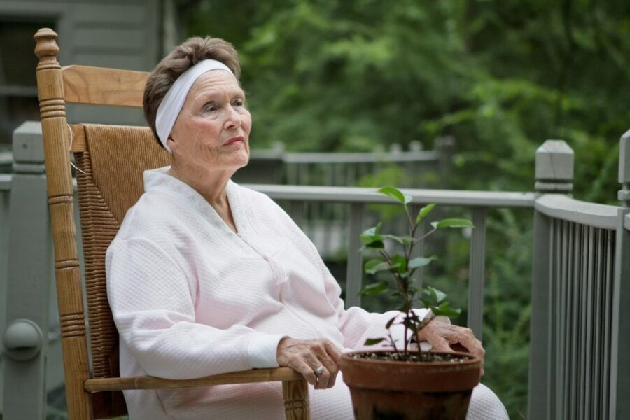 Older woman in white headband and pink robe sits in wooden rocking chair with potted plant on outdoor deck, Final Acts