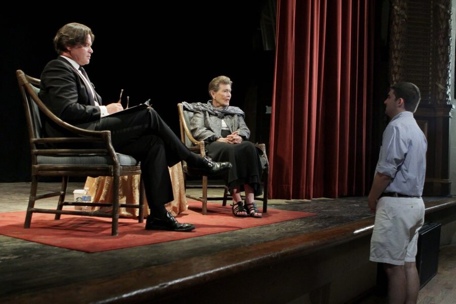 Director observing two people seated in chairs during a stage panel discussion with red curtain backdrop