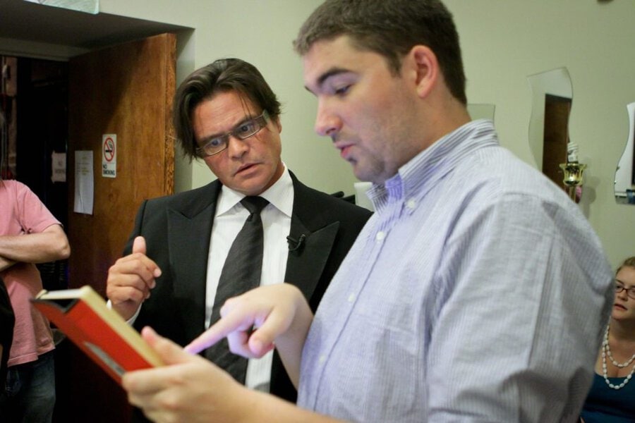 Man in black suit with glasses and man in blue striped shirt review script pages together on film set