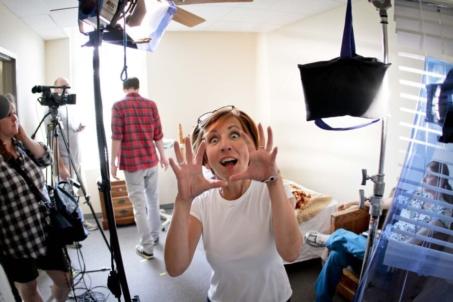 Woman in white shirt making surprised expression with raised hands on film set with crew and lighting equipment