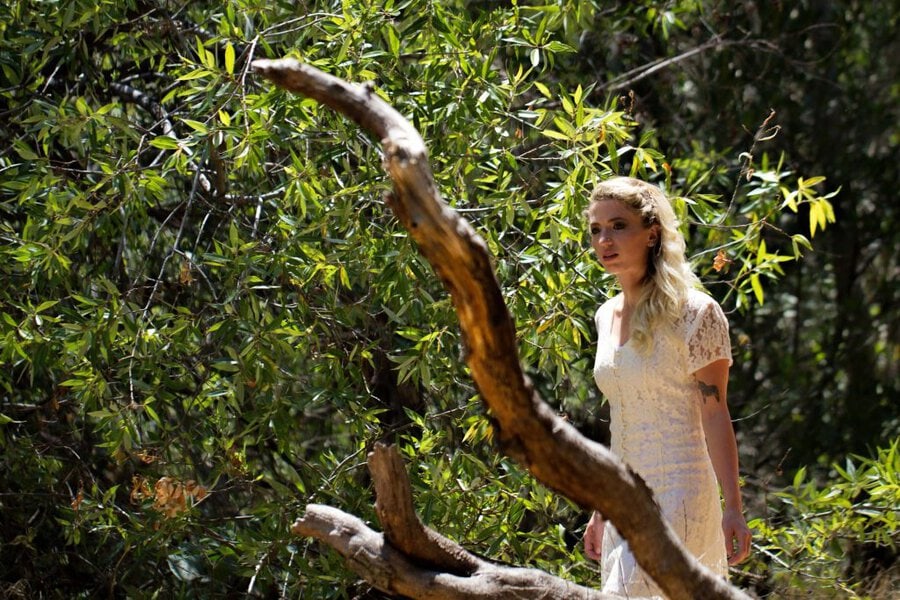 Woman in white lace dress standing among twisted branches in sunlit forest during music video shoot
