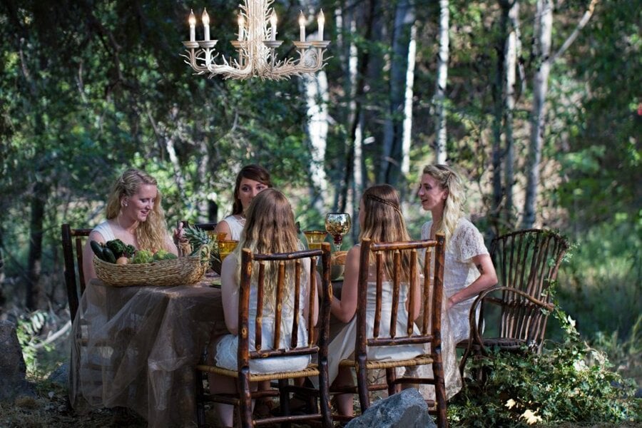 Five women in white dresses seated at rustic table under antler chandelier in forest, Branches music video (2015)