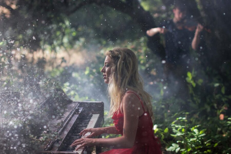 Woman in red dress playing keyboard outdoors as water droplets spray around her, forest background, Branches music video