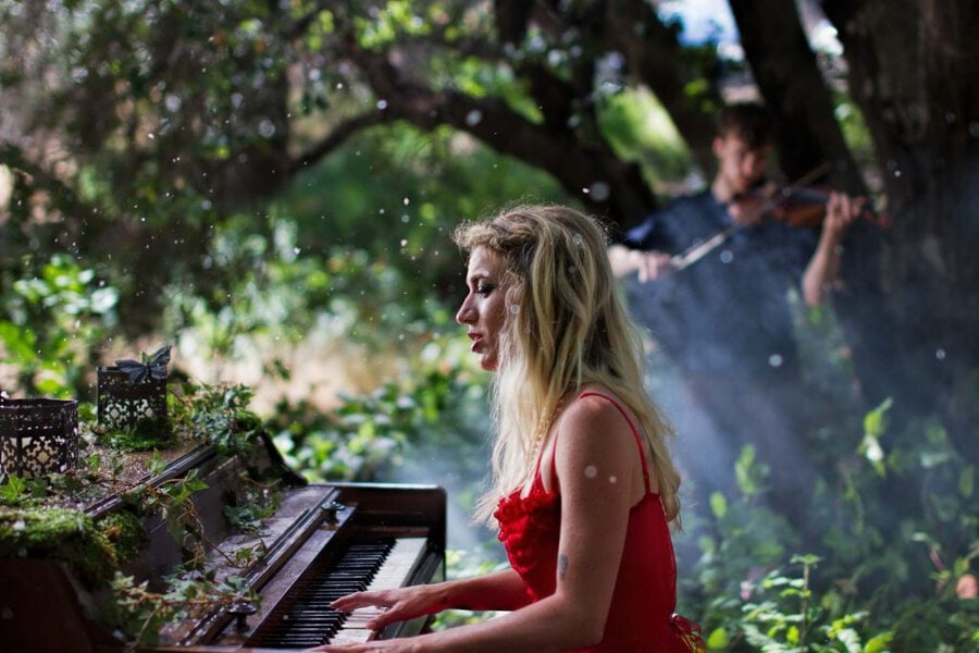 Blonde pianist in red dress plays overgrown piano in forest setting, violinist stands behind, sunlight filters through trees