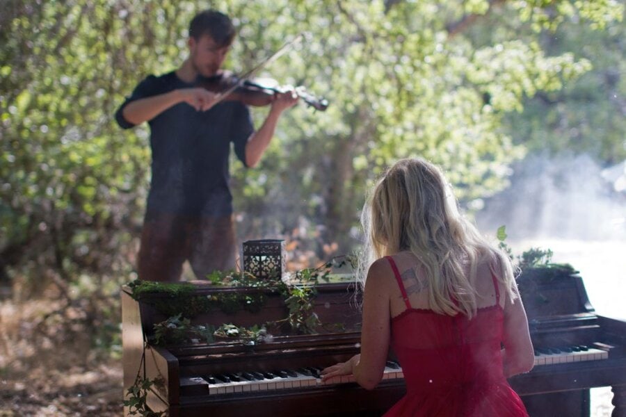 A violinist and pianist perform together outdoors on a moss-covered piano in a sunlit forest setting