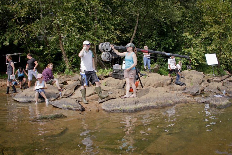 Film crew with camera jib on rocks beside brown river, surrounded by trees during outdoor shoot in Macon, GA