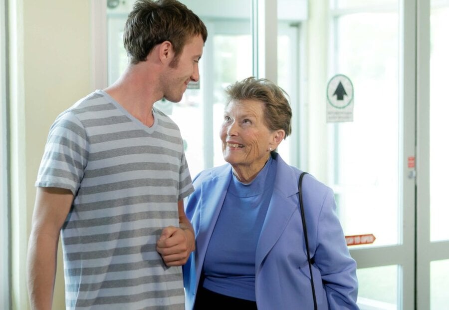 Young man in striped shirt walking arm-in-arm with older woman in blue jacket in bright hallway