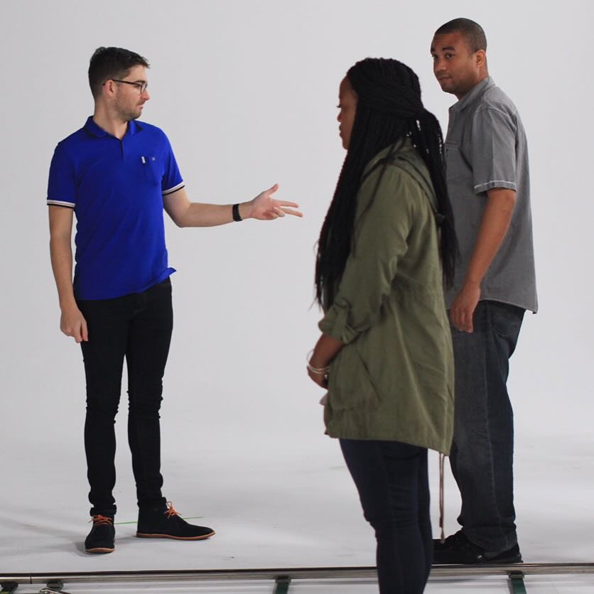 Director in blue polo gestures while speaking to two models on white studio set for We the Future commercial shoot