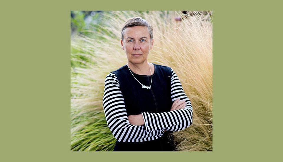 Person with short hair and a striped shirt stands confidently with arms crossed in front of tall, wispy grass, against a green background.