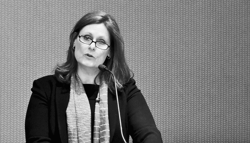A woman with glasses speaks into a microphone. She wears a dark blazer and a striped scarf. The background is a textured wall. The image is in black and white.