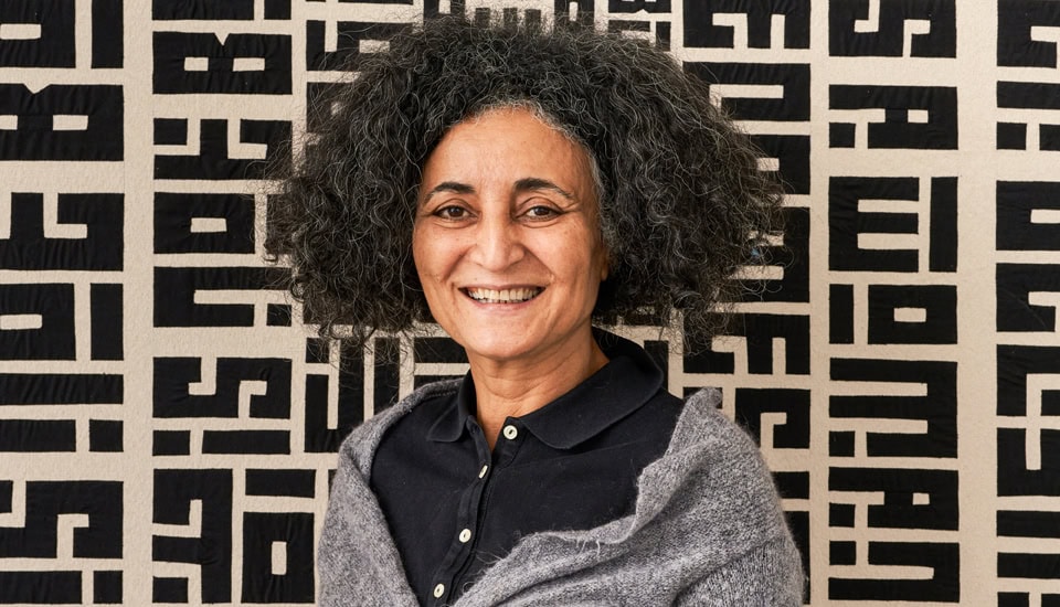 A person with curly hair smiles while standing in front of a patterned black and white background. They are wearing a dark collared shirt and a gray shawl. The background features bold geometric text designs.