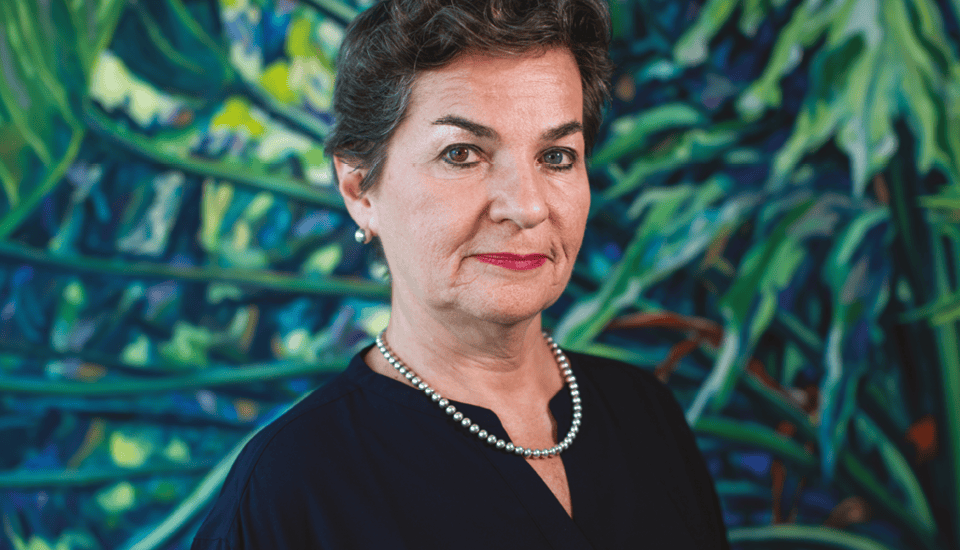 Woman with short hair and pearl necklace stands against a vibrant, abstract green and blue background, wearing a black top.