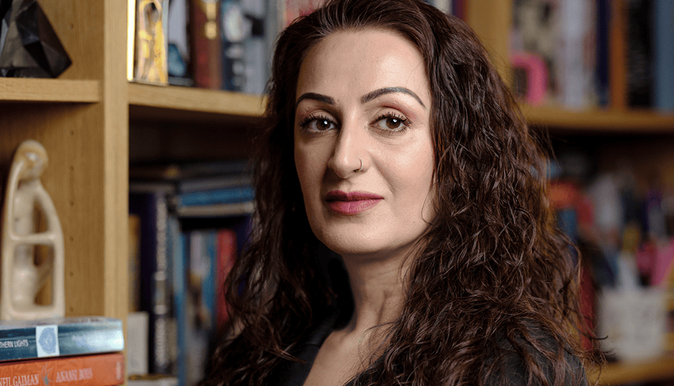 A person with long, curly hair and a nose ring stands in front of a bookcase, looking toward the camera. The background is filled with books and a small sculpture.