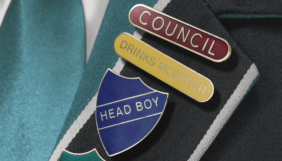 Close-up of a school blazer showing three badges: a red "Council" badge, a yellow "Drinks Monitor" badge, and a blue shield-shaped "Head Boy" badge. A teal tie is visible underneath the blazer.