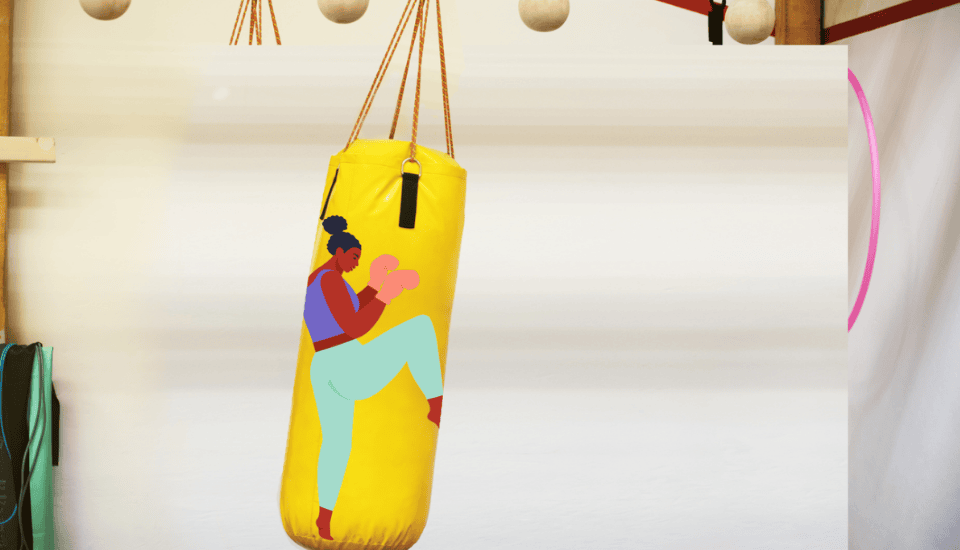A vibrant yellow punching bag hangs from the ceiling in a gym. It is decorated with an illustration of a person in a boxing pose wearing mint green pants and a purple top. Other gym equipment is visible in the background.