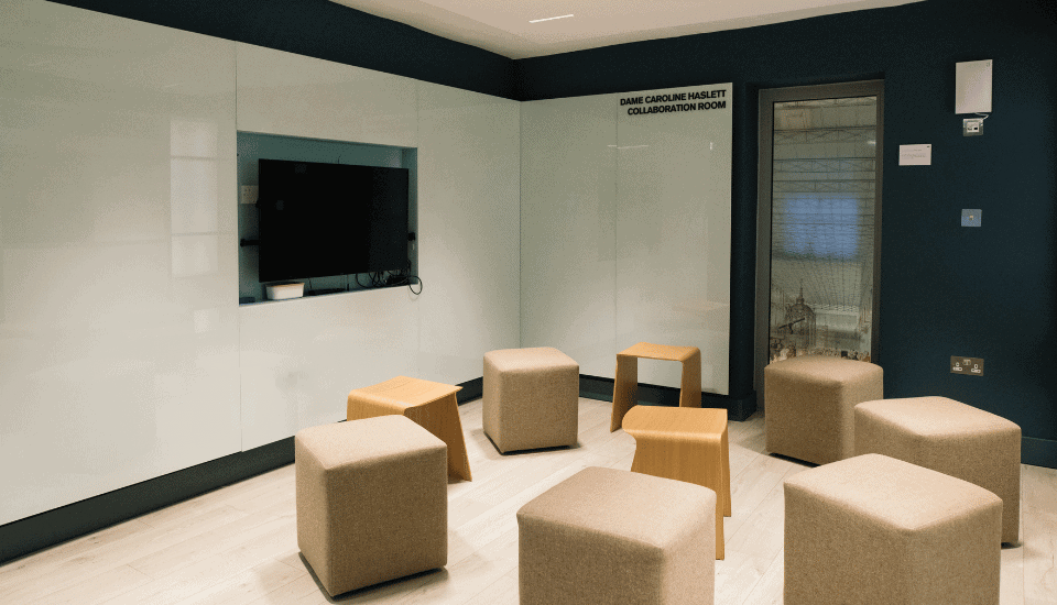 A modern meeting room with a large screen mounted on a whiteboard wall. Beige cube and hexagonal stools are arranged randomly on a light wood floor. A door with a glass panel is on the side. The room is lit with recessed lighting.