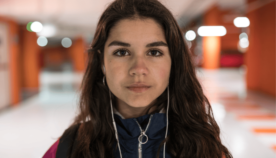 A young woman with long dark hair stands in a brightly lit hallway, wearing a jacket and earphones. The background is slightly blurred, with orange and white walls. She looks directly at the camera with a neutral expression.