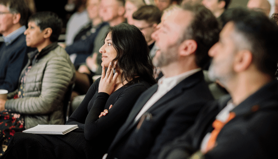 A diverse group of people seated in an audience. A woman in the foreground, wearing a black top, looks attentive with her chin resting on her hand. Others around her are engaged, some smiling. The setting appears to be a lecture or presentation.