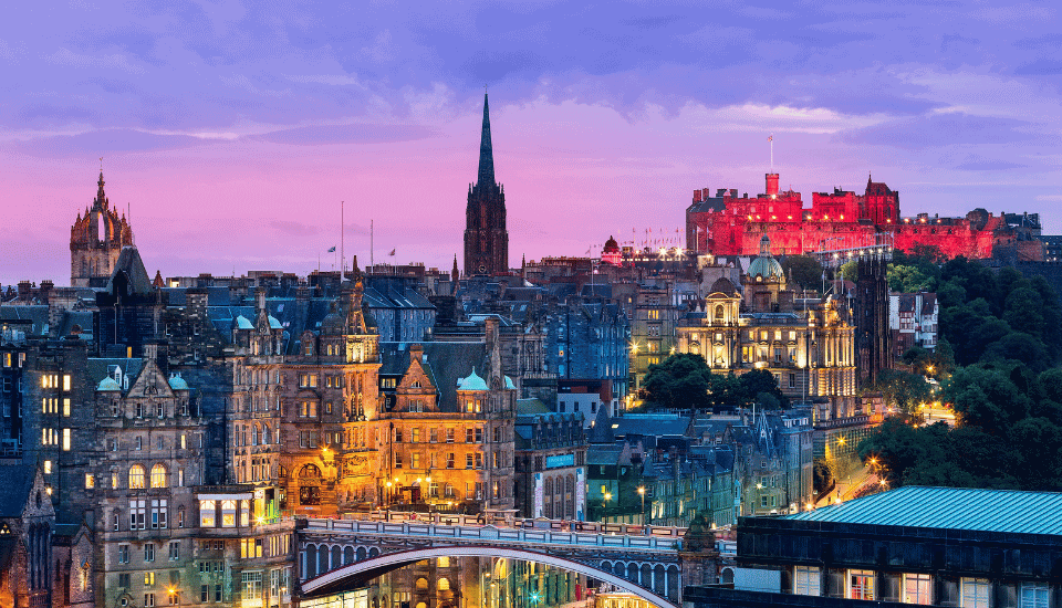 Skyline of a city at twilight with historical buildings. A spire rises prominently in the center, and a castle is visible on a hilltop to the right, illuminated with red light. The sky is a gradient of pink and purple hues.