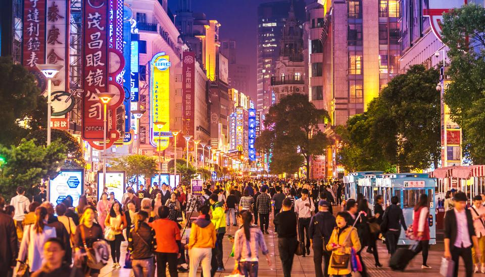 A bustling night scene of a vibrant city street filled with people walking. Bright, colorful signs in various languages illuminate the street, with tall buildings lining both sides. The atmosphere is lively and energetic.