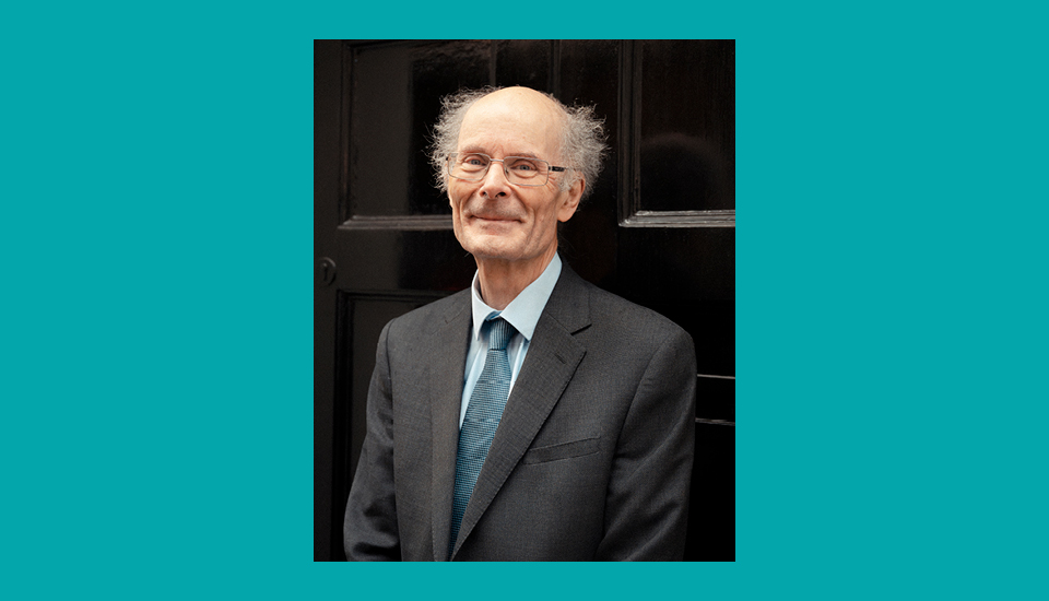 Elderly man with glasses and thinning hair wearing a suit and tie, standing in front of a dark door, smiling faintly.