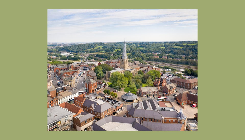 Aerial view of an East Midlands town with a prominent spire rising from a church surrounded by trees. Historic brick buildings and narrow streets weave through the landscape, while rolling green hills stretch into the distance under a partly cloudy sky, embodying inclusive growth.