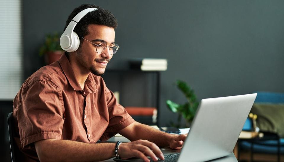 A person wearing glasses and headphones sits at a desk, smiling while using a laptop. The background features a plant and shelves with books against a dark wall.