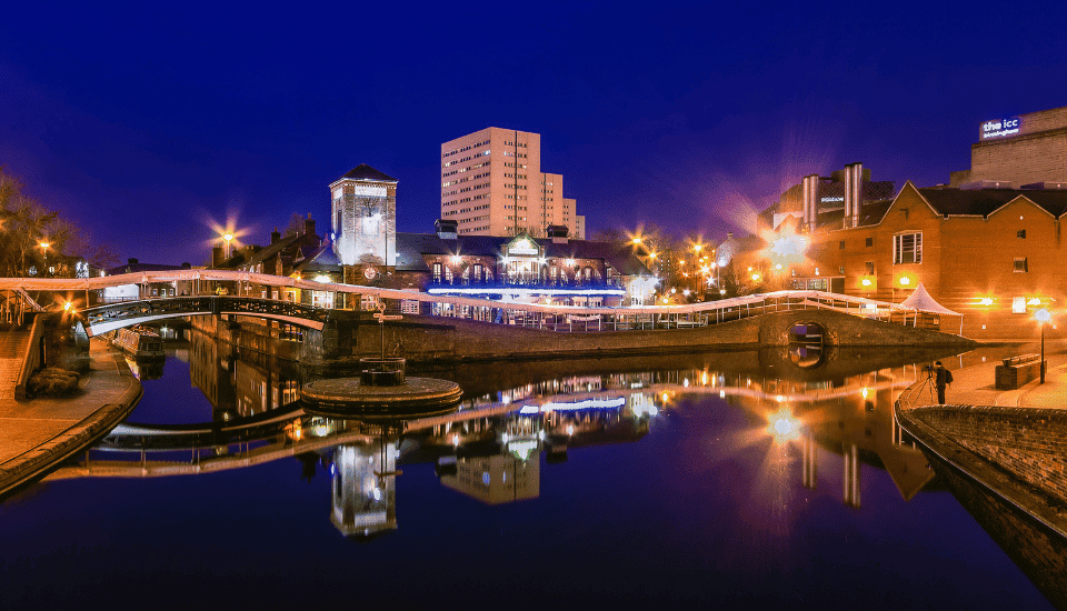 Night view of a canal in a city featuring bridges and illuminated buildings symbolizing inclusive growth, with their bright reflections shimmering on the calm water beneath a deep blue sky.