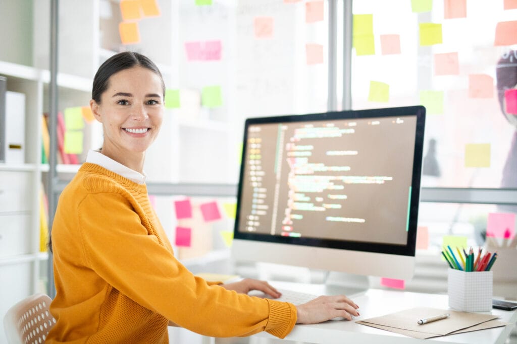 Website erstellen: Eine hübsche junge Webdesignerin in legerer Kleidung schaut Sie von ihrem Schreibtisch mit dem Computer aus an, während sie an einem neuen Online-Shop-Projekt arbeitet.