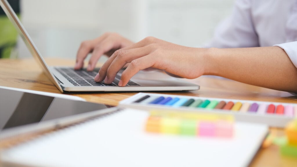 Das Foto zeigt eine Nahaufnahme der Hände einer Person, die auf der Tastatur eines Laptops tippen. Der Laptop steht auf einem Holztisch, der Rand des Bildschirms ist heruntergeklappt, so dass der Bildschirminhalt nicht sichtbar ist. Die Person scheint ein weißes Hemd oder eine weiße Bluse zu tragen, von der im Bild nur die Ärmel zu sehen sind. Neben dem Laptop liegt ein Spiralnotizbuch mit einem Satz bunter Pastell- oder Filzstifte. Die Stifte sind in verschiedenen Farben erhältlich, darunter grün, blau, lila, rot und andere. Das Gesamtbild erinnert an einen kreativen Arbeitsplatz oder möglicherweise an eine Design- oder Kunstumgebung. Die Szene vermittelt ein Gefühl von Produktivität und Organisation.