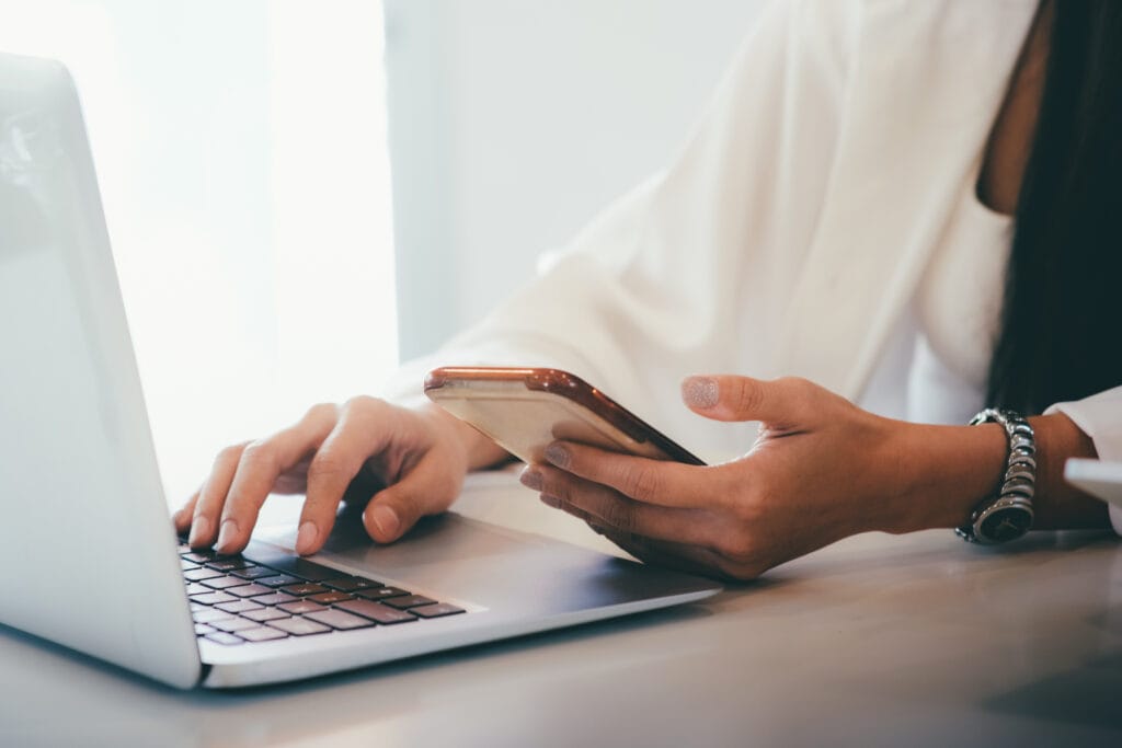 Search Generative Experience (SGE). Das Foto zeigt eine Nahaufnahme der Hände einer Person, die mit zwei elektronischen Geräten interagiert. Eine Hand tippt leicht auf der Tastatur eines Laptops, während die andere ein Smartphone hält. Der Laptop steht auf einer Oberfläche, möglicherweise einem Tisch, aber der konkrete Inhalt des Bildschirms ist nicht zu erkennen. Die Person scheint eine weiße Bluse oder ein weißes Hemd zu tragen. Einer ihrer Fingernägel ist mit glitzerndem Nagellack verziert. Außerdem trägt sie ein Armband, das aus mehreren Bändern besteht und dadurch mehrschichtig wirkt. Das Licht auf dem Foto ist weich und beleuchtet die Szene sanft. Die Gesamtatmosphäre deutet auf einen modernen Arbeitsplatz oder eine Umgebung hin, in der Multitasking vorherrscht, was die Anforderungen des digitalen Zeitalters widerspiegeln könnte.