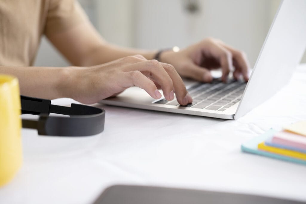 Das Foto zeigt eine Nahaufnahme der Hände einer Person, die auf der Tastatur eines Laptops tippt. Die Kleidung oder das Oberteil der Person ist nicht vollständig zu erkennen, scheint jedoch beige oder hellbraun zu sein. Im Vordergrund liegt ein schwarzer Kopfhörer oder ein Headset auf einem Tisch und ein gelber Gegenstand, möglicherweise eine Tasse oder ein Becher, ist teilweise sichtbar. In der Nähe des Laptops befinden sich Stapel verschiedenfarbiger Haftnotizen. Die Umgebung scheint ein ruhiger und organisierter Arbeitsplatz mit einer hellen Tischoberfläche zu sein. Der Bildschirm des Laptops ist angewinkelt, so dass sein Inhalt in der Aufnahme nicht sichtbar ist. Die Gesamtsituation lässt auf eine konzentrierte Arbeits- oder Lernumgebung schließen.