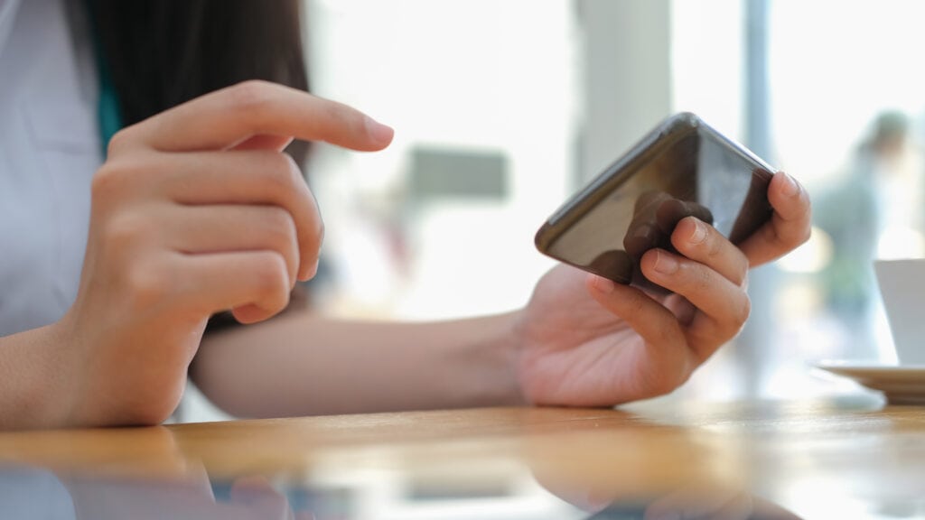 Google Search Generative Experience (SGE). Das Foto zeigt eine Nahaufnahme der Hände einer Person, die mit einem Smartphone interagiert. Eine Hand hält das Telefon fest, während der Zeigefinger der anderen Hand vermutlich zum Tippen oder Wischen auf dem Bildschirm positioniert ist. Der Hintergrund ist unscharf, deutet aber auf einen Innenraum mit natürlichem Lichteinfall hin, möglicherweise durch ein Fenster. Auf einem Tisch oder einer Arbeitsplatte ist die schwache Silhouette eines anderen Gegenstandes, möglicherweise einer Tasse oder eines Bechers, zu erkennen. Die Person scheint ein weißes oder helles Oberteil zu tragen, obwohl nur ein kleiner Teil davon sichtbar ist. Der Schwerpunkt des Bildes liegt auf den Händen, dem Telefon und der Tätigkeit, die gerade ausgeführt wird, wodurch ein Moment der digitalen Beschäftigung oder Kommunikation vermittelt wird. Die gesamte Atmosphäre ist ruhig und entspannt, was auf eine zwanglose Umgebung schließen lässt.