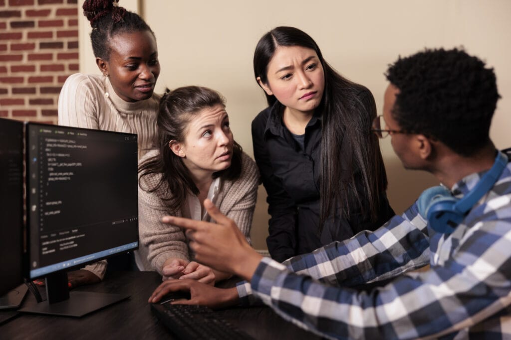 Auf dem Foto ist ein Team von vier Entwicklern zu sehen, die sich aufmerksam über einen Arbeitsplatz mit Computermonitoren beugen. Ein Mann im Vordergrund, der ein kariertes Hemd trägt und Kopfhörer um den Hals hat, scheint seinen Kolleginnen etwas auf dem Bildschirm zu zeigen oder zu erklären. Drei Frauen hören ihm zu und betrachten interessiert den Bildschirm, wobei ihre Gesichtsausdrücke Konzentration und Nachdenklichkeit vermuten lassen. Eine der Frauen zeigt auf den Bildschirm, was darauf hindeutet, dass sie eine Frage oder einen Kommentar zu dem hat, was sie sehen. Der Bildschirm zeigt Programmcode, was nahelegt, dass sie an einer komplexen Aufgabe wie der Entwicklung einer maschinellen Lernanwendung mit Amazon SageMaker arbeiten könnten. Der Raum hat eine warme, einladende Atmosphäre mit Ziegelwand im Hintergrund.