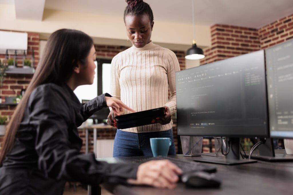 Auf dem Foto ist ein Büro zu sehen, in dem zwei Entwicklerinnen zusammenarbeiten. Eine der Entwicklerinnen sitzt links im Bild und trägt eine schwarze Bluse; sie zeigt auf etwas auf dem Bildschirm oder auf ein Gerät, das von der zweiten Entwicklerin gehalten wird. Diese steht rechts im Bild, trägt einen beige Pullover und Jeans und blickt auf das Gerät in ihrer Hand, das einem Tablet ähnelt. Vor ihnen sind zwei Computermonitore, auf denen Code zu sehen ist. Dies könnte darauf hindeuten, dass sie an einem Projekt im Bereich des maschinellen Lernens arbeiten, möglicherweise mit einem Tool wie Amazon SageMaker. Das Büro ist modern gestaltet mit einer Ziegelwand im Hintergrund und einer Pflanze auf dem Schreibtisch, was auf eine angenehme Arbeitsatmosphäre hindeutet.
